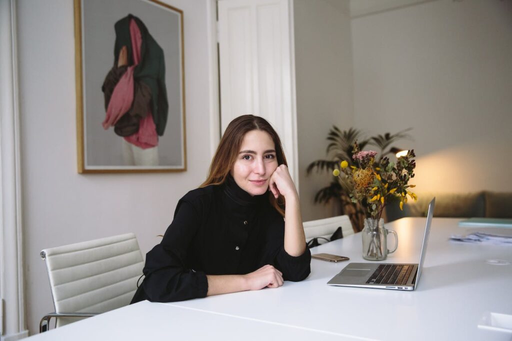 dziewczyna w czarnym swetrze, siedząca na przeciwko laptopa uśmiecha się w kierunku kamery